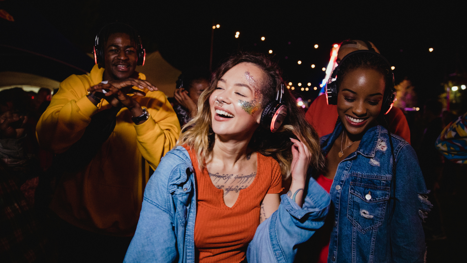 A group of young people with glitter on their faces dance happily in a dark outdoor location with headphones on